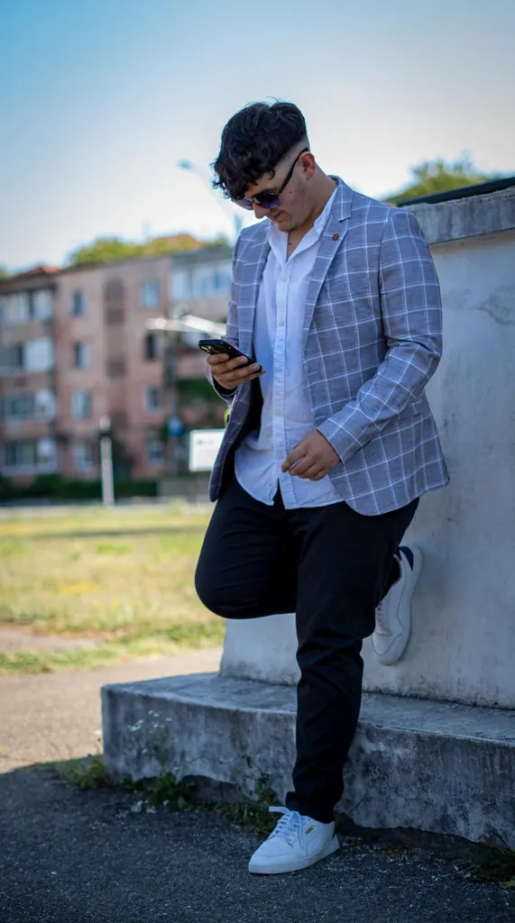 Tânăr în sacou în carouri, sprijinit de un zid, cu telefonul în mână