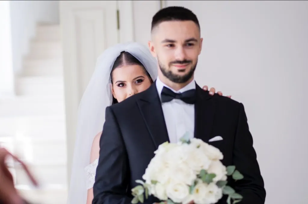 Mireasa privește jucăuș din spatele mirelui, care ține buchetul de trandafiri albi